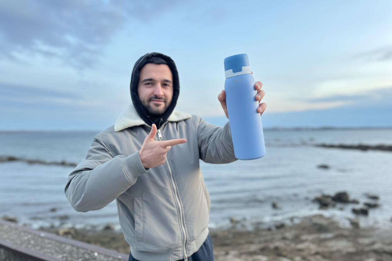 Person holding a blue SafeSip water filtered bottle by the ocean, blue sky and great photography. Stay hydrated with NOMAD