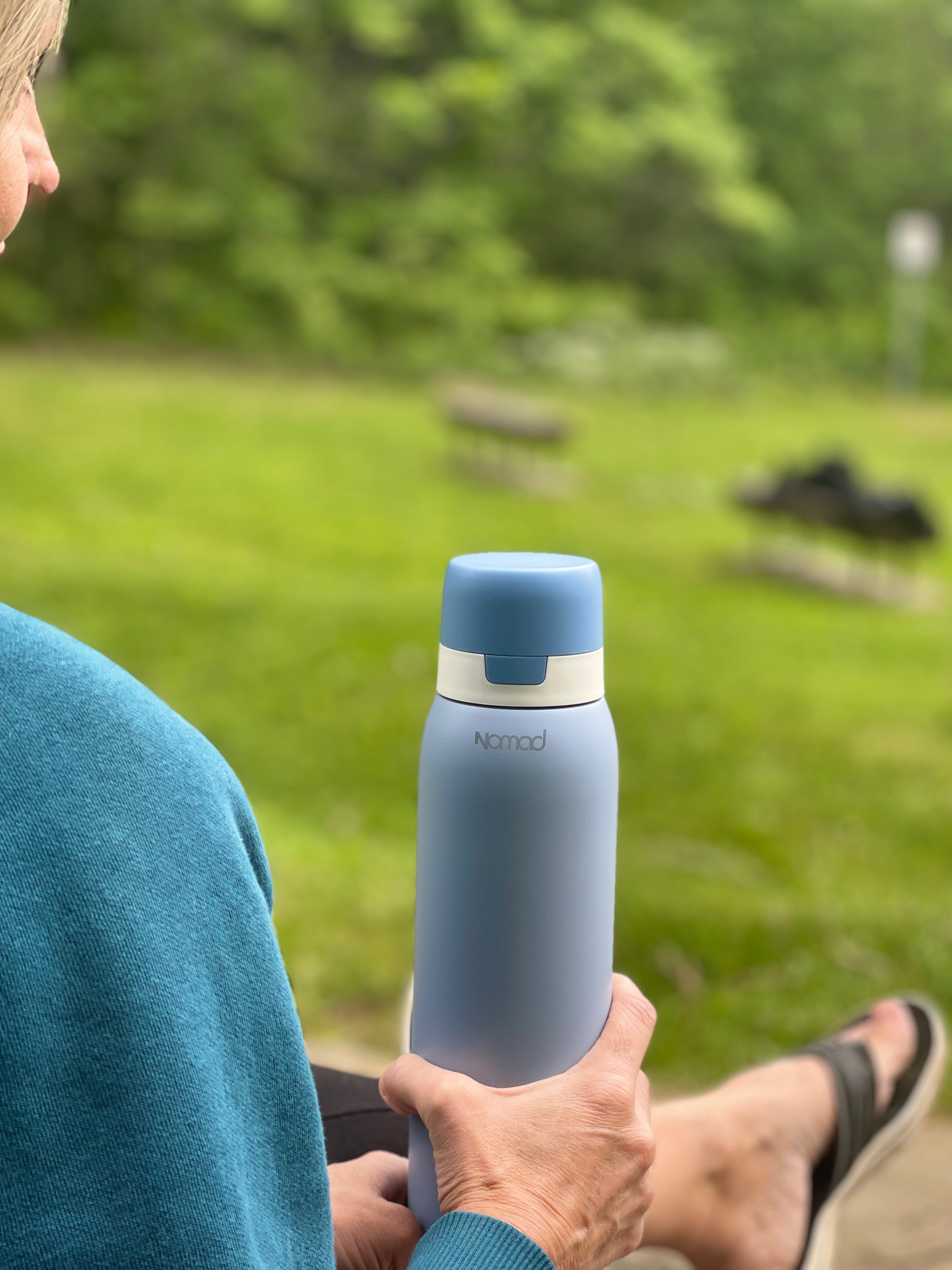 Person holding a blue and gray insulated NOMAD bottle outdoors with greenery in the background