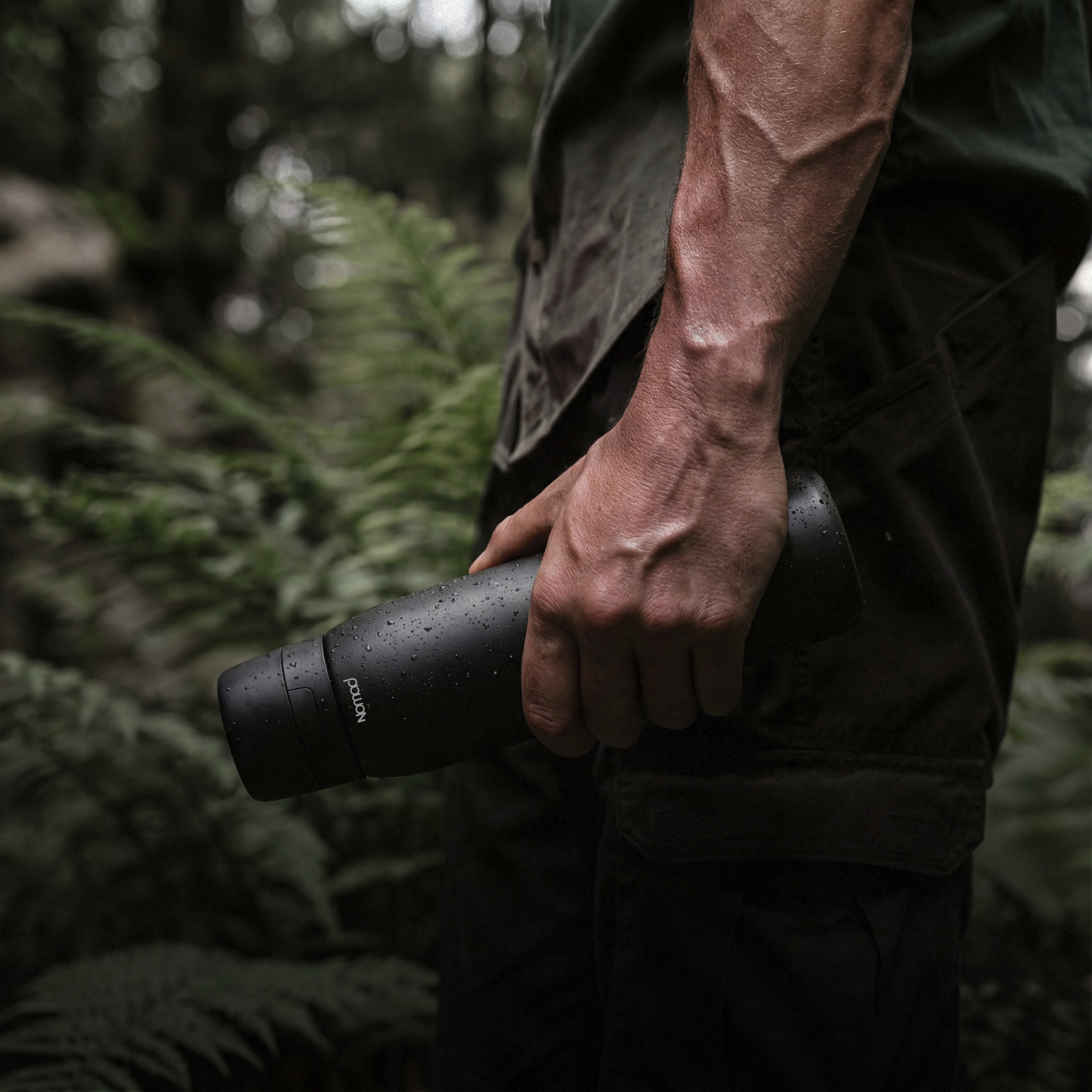 Hand holding a black NOMAD insulated water bottle in a forest, outdoor adventure lifestyle product image with cinematic lighting