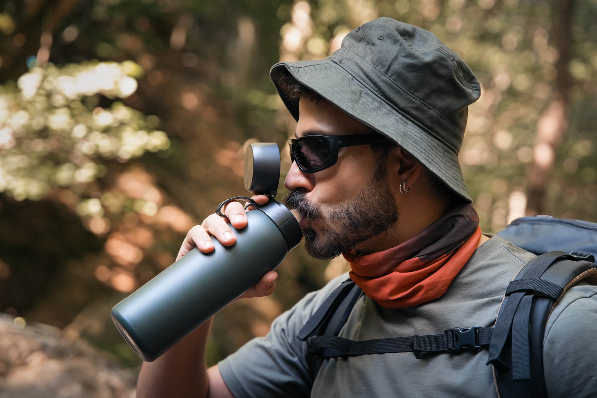 A man wearing a green bucket hat, sunglasses, and a gray t-shirt drinks from a matte dark green insulated NOMAD SafeSip water purifier bottle with a black lid in a forest setting.