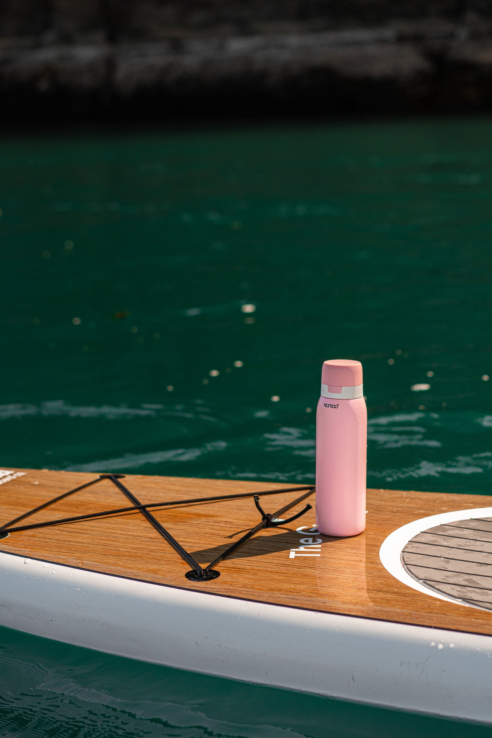A pink insulated NOMAD SafeSip water bottle with a matching pink cap stands on the deck of a paddleboard on green water.