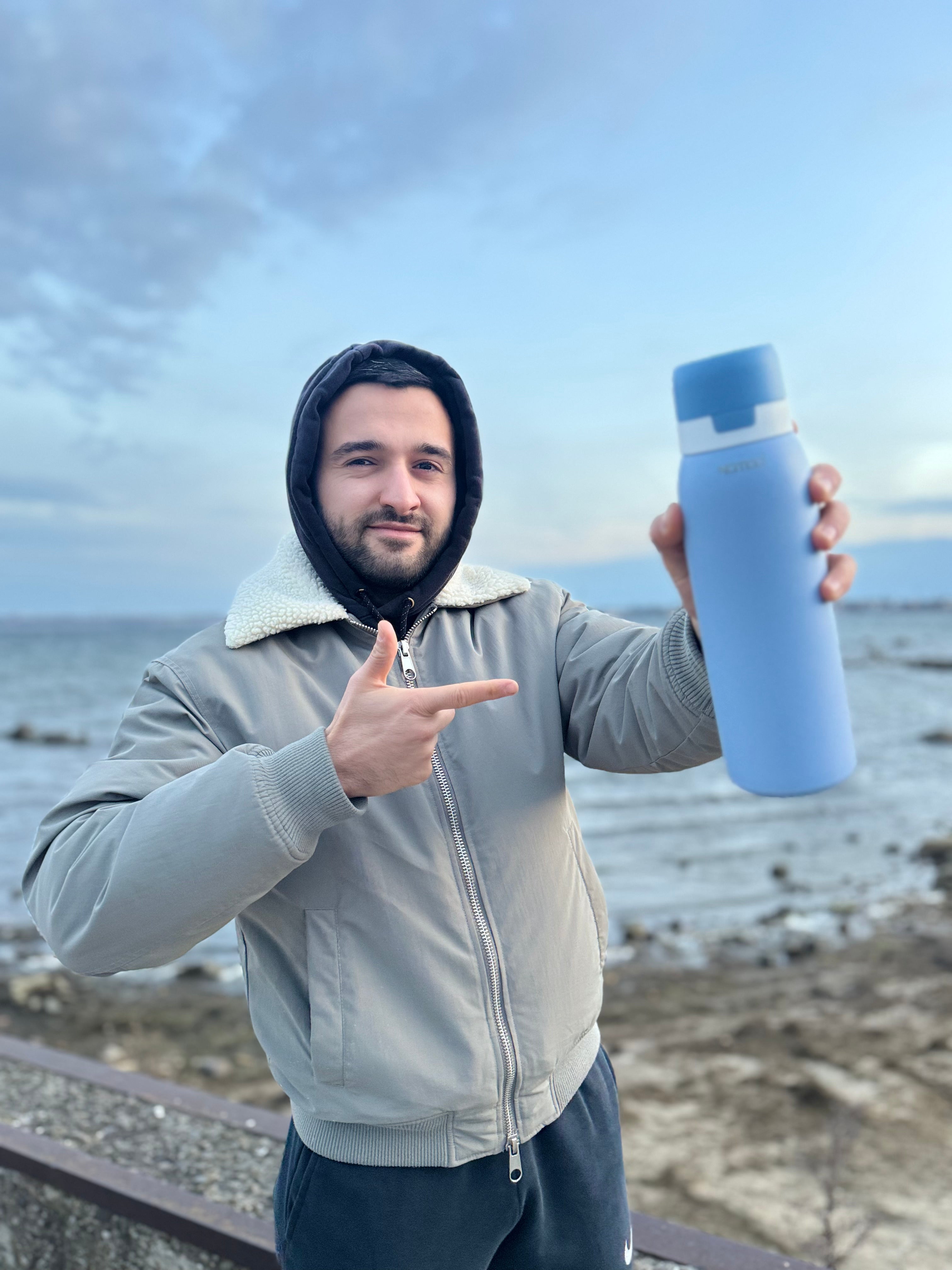 Person holding a blue SafeSip water filtered bottle by the ocean, blue sky and great photography. Stay hydrated with NOMAD