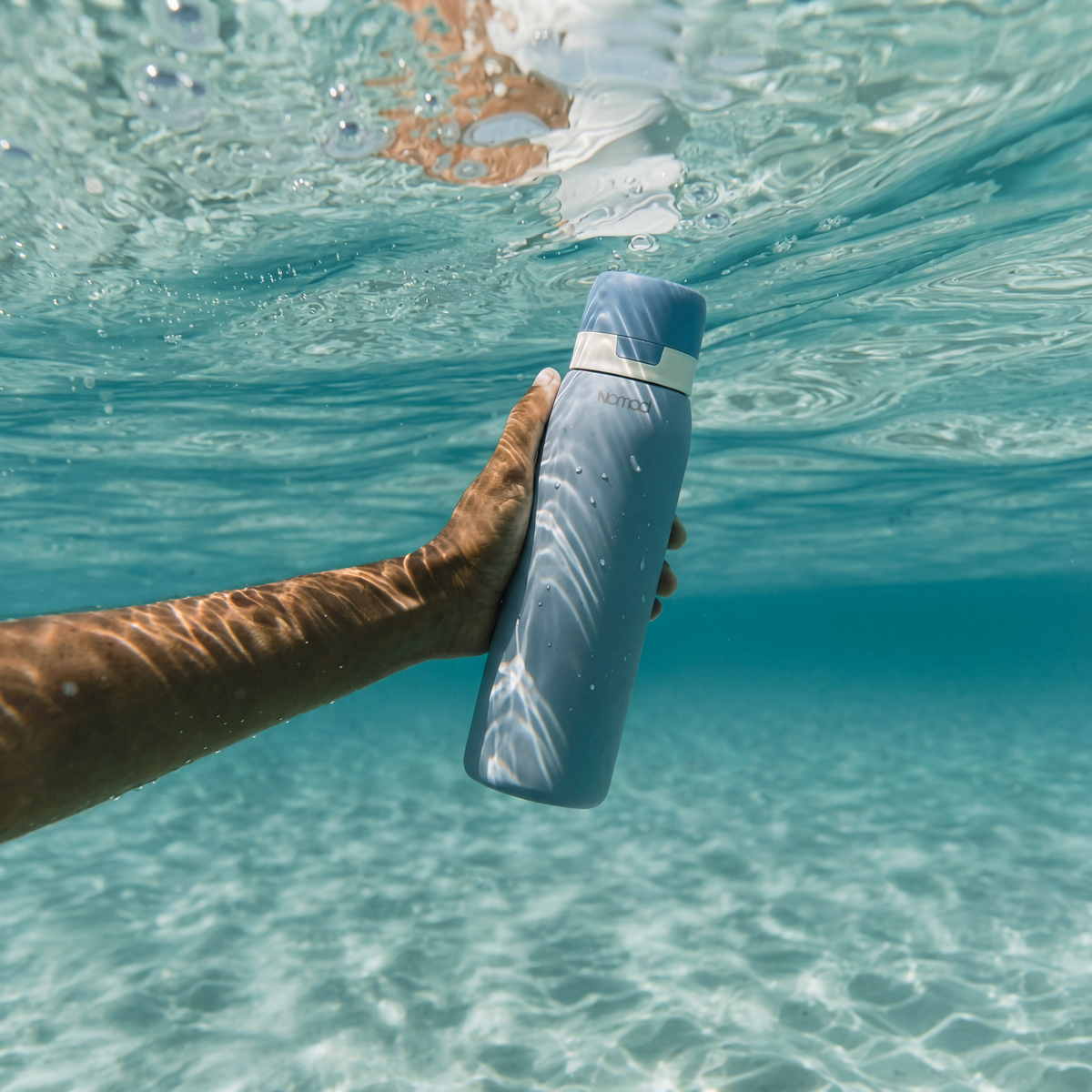 Hand holding a blue NOMAD water bottle underwater with clear blue water in the background