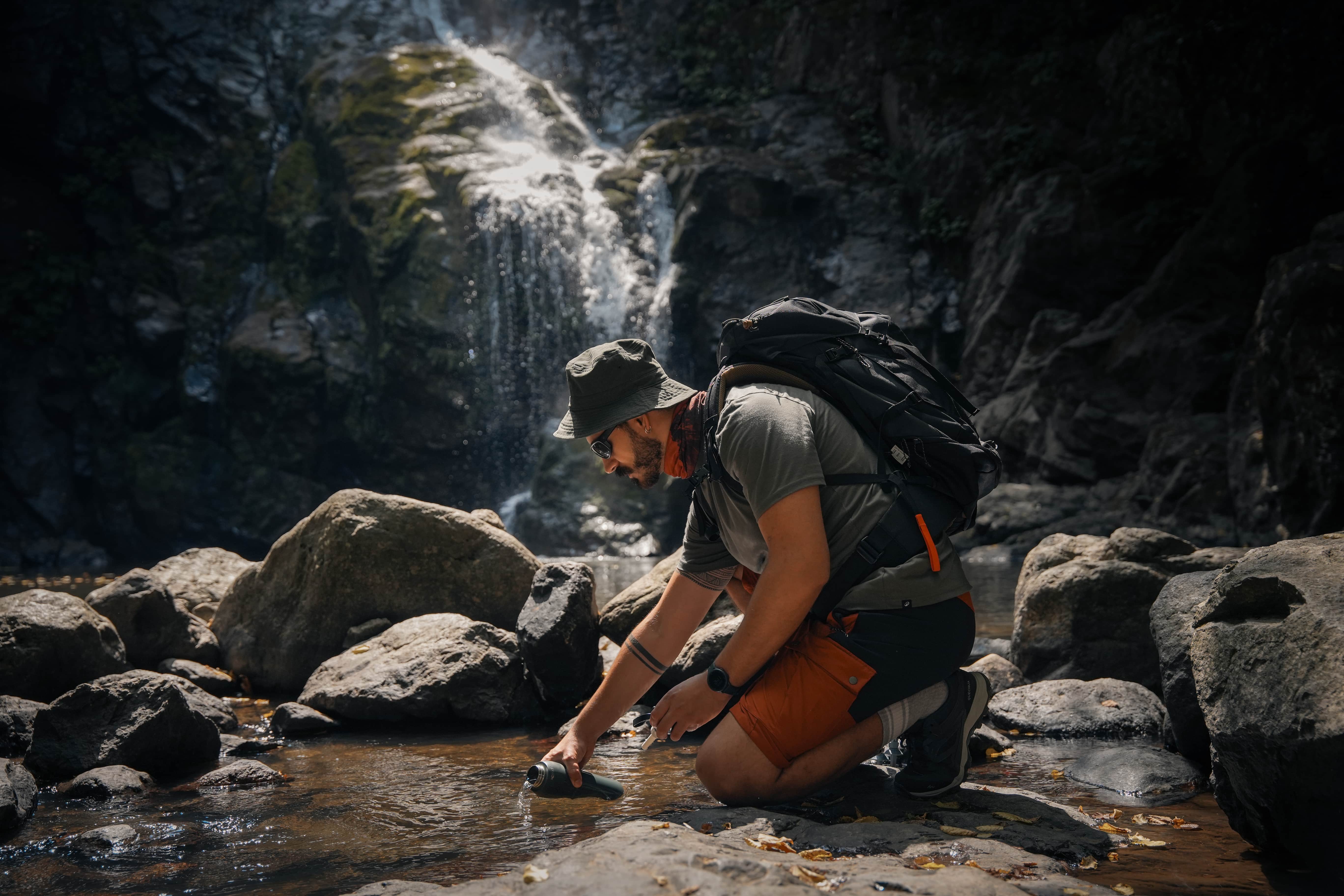 A hiker kneels beside a rocky stream to fill a Forest Green NOMAD SafeSip portable water purifier bottle, surrounded by a forest canyon and waterfall during an outdoor adventure.