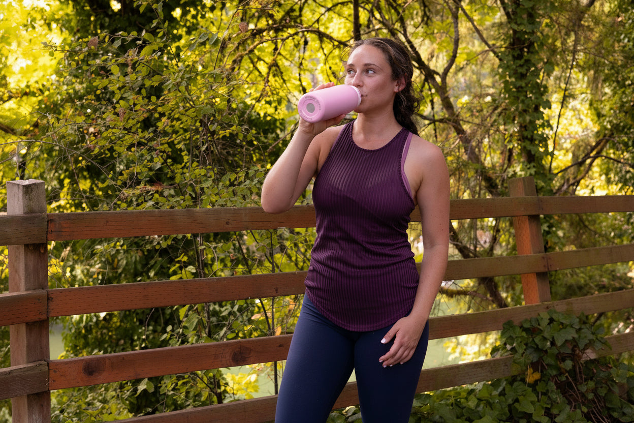 Woman in athletic wear drinking from a pink NOMAD reusable bottle on a wooden bridge in a lush forest, capturing real outdoor hydration on an adventure.