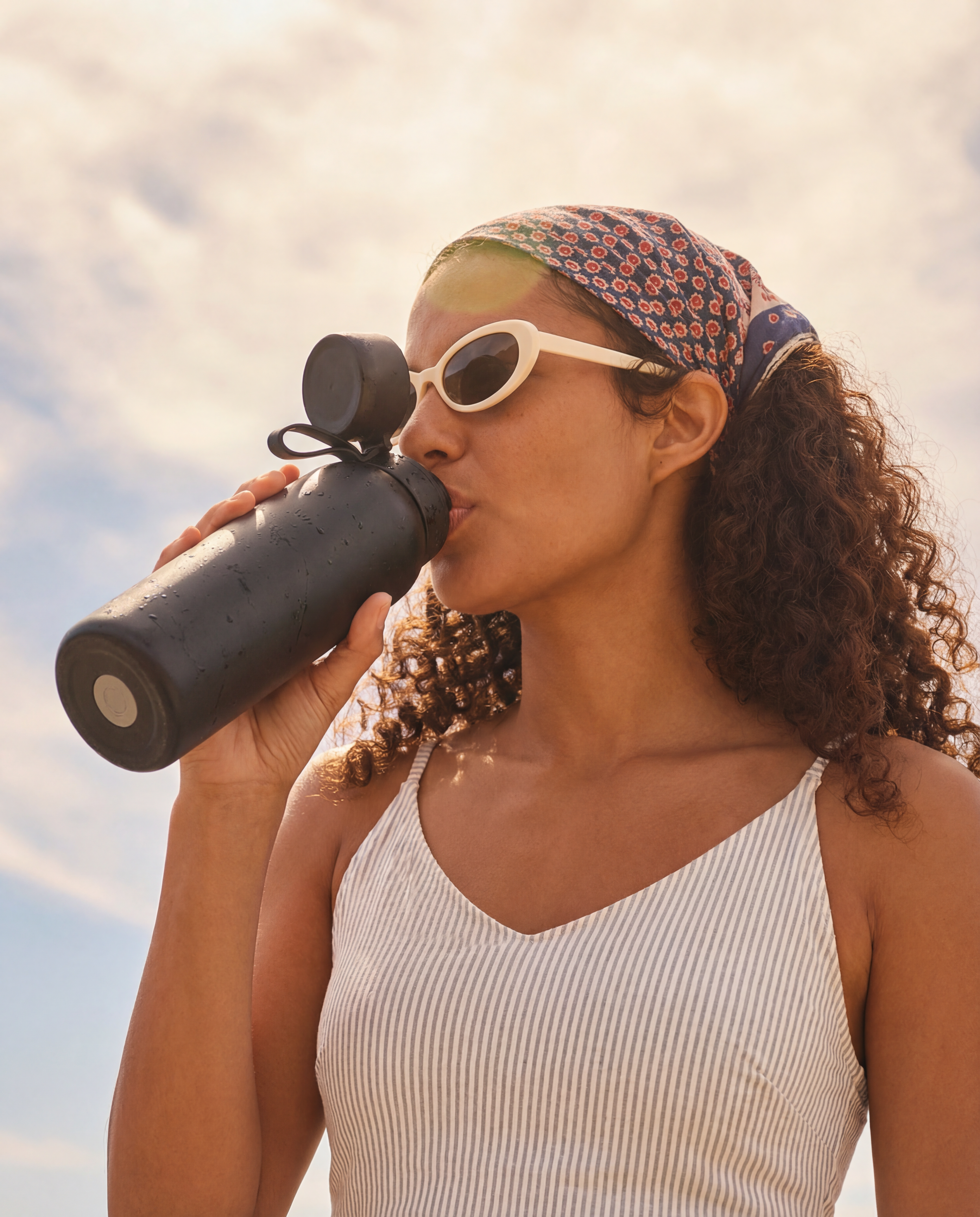 Woman drinking from a NOMAD green forest filtered water bottle with a scenic sky background