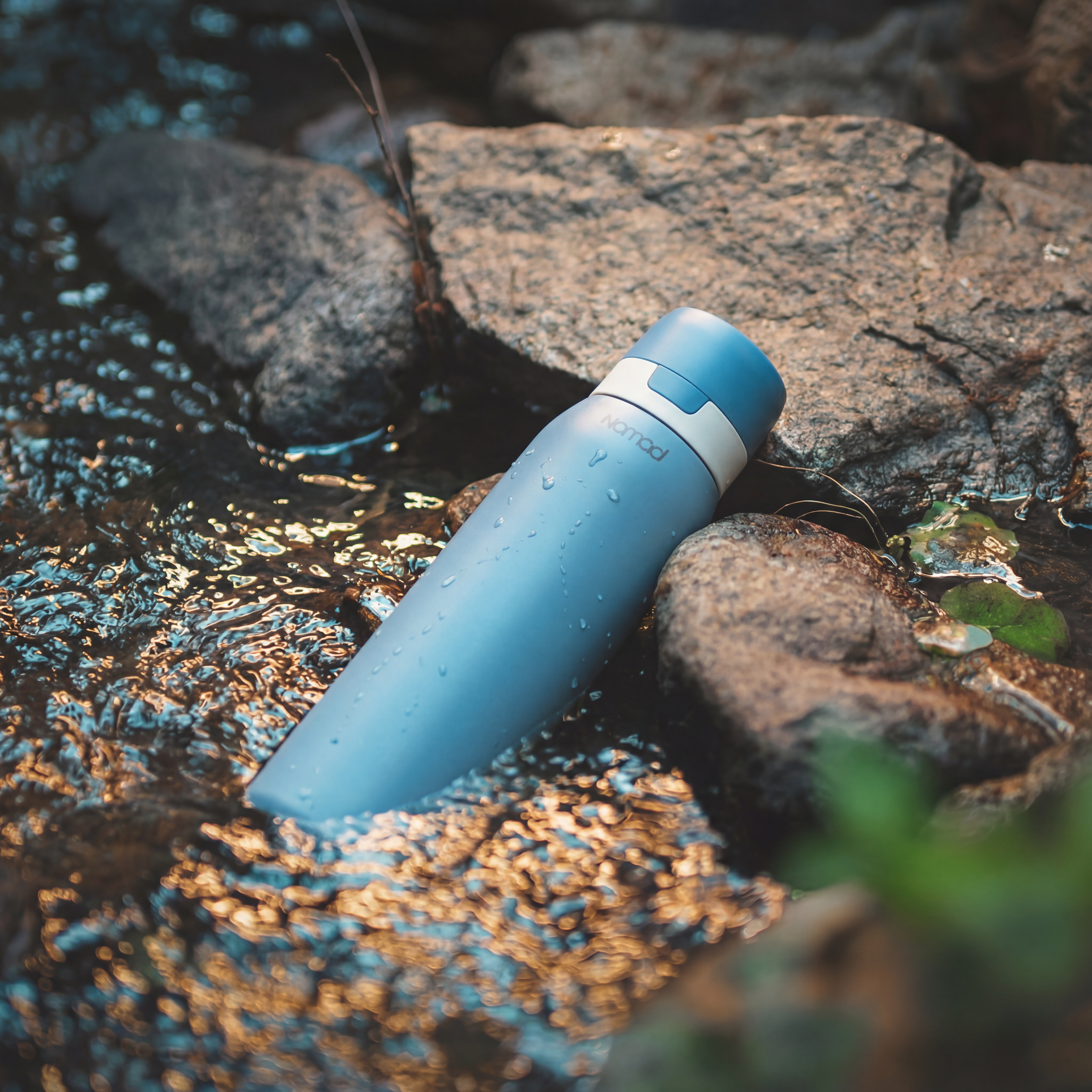 NOMAD SafeSip reusable filtered water bottle resting in a clear stream among rocks, designed for outdoor adventures, hiking, travel, and everyday hydration.