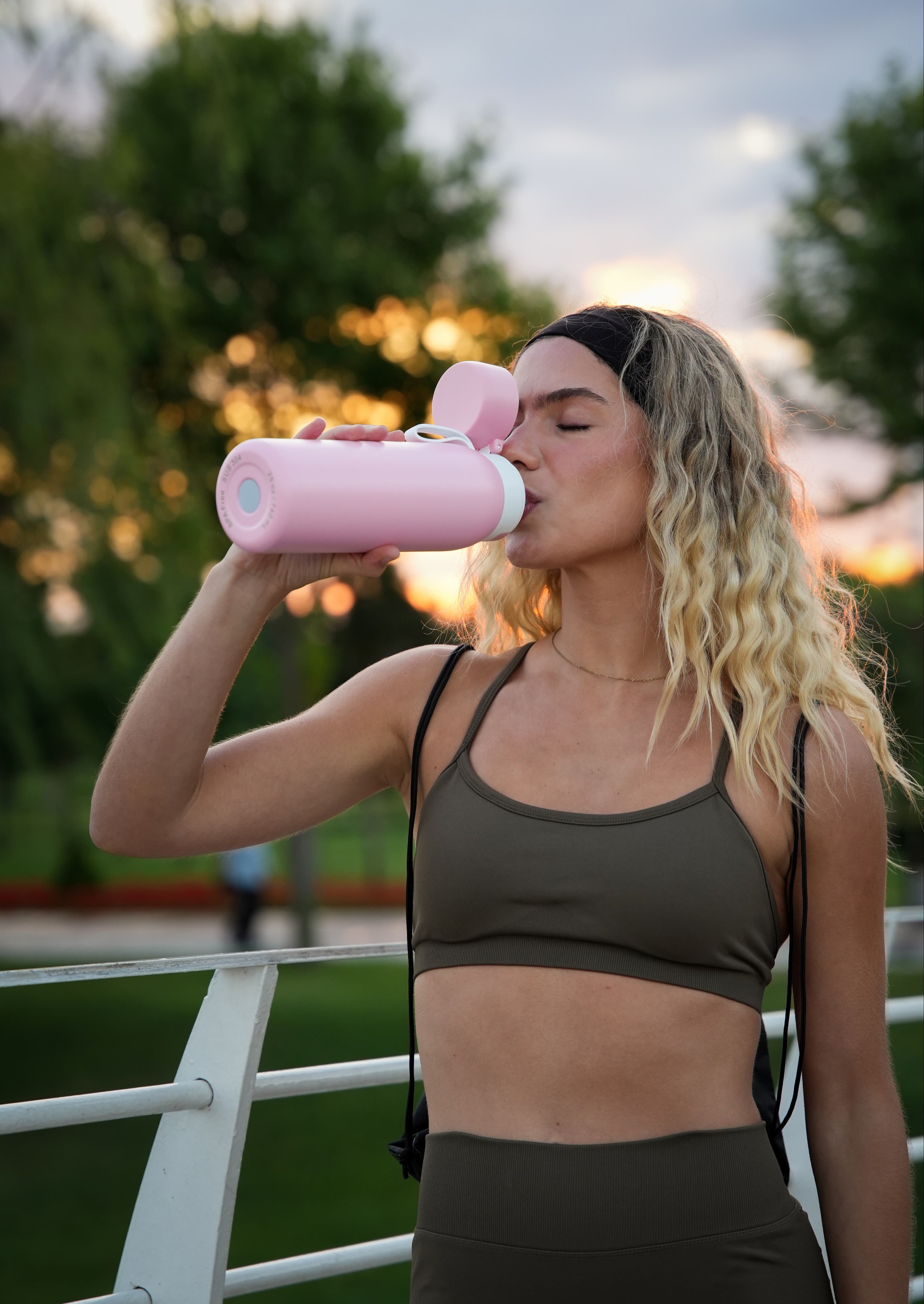 Woman runner drinking from a pink NOMAD SafeSip insulated purifier bottle during an outdoor run at sunset.