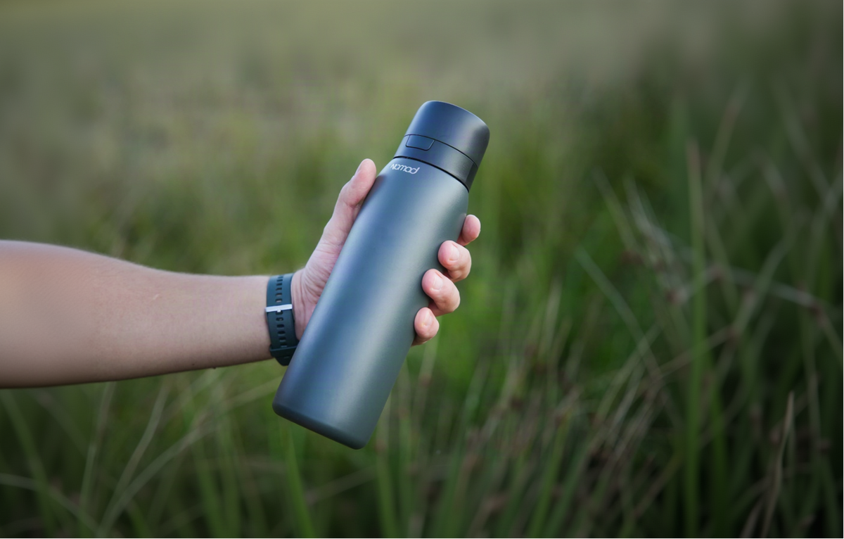 A person's hand holding a matte dark gray NOMAD water bottle outdoors against a blurred green grassy background.