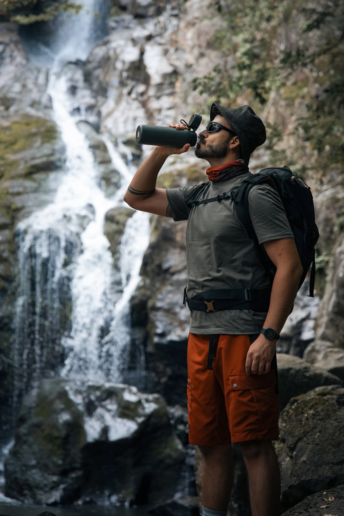 Hiker drinking from a black NOMAD SafeSip water purifier bottle while resting on a forest trail.