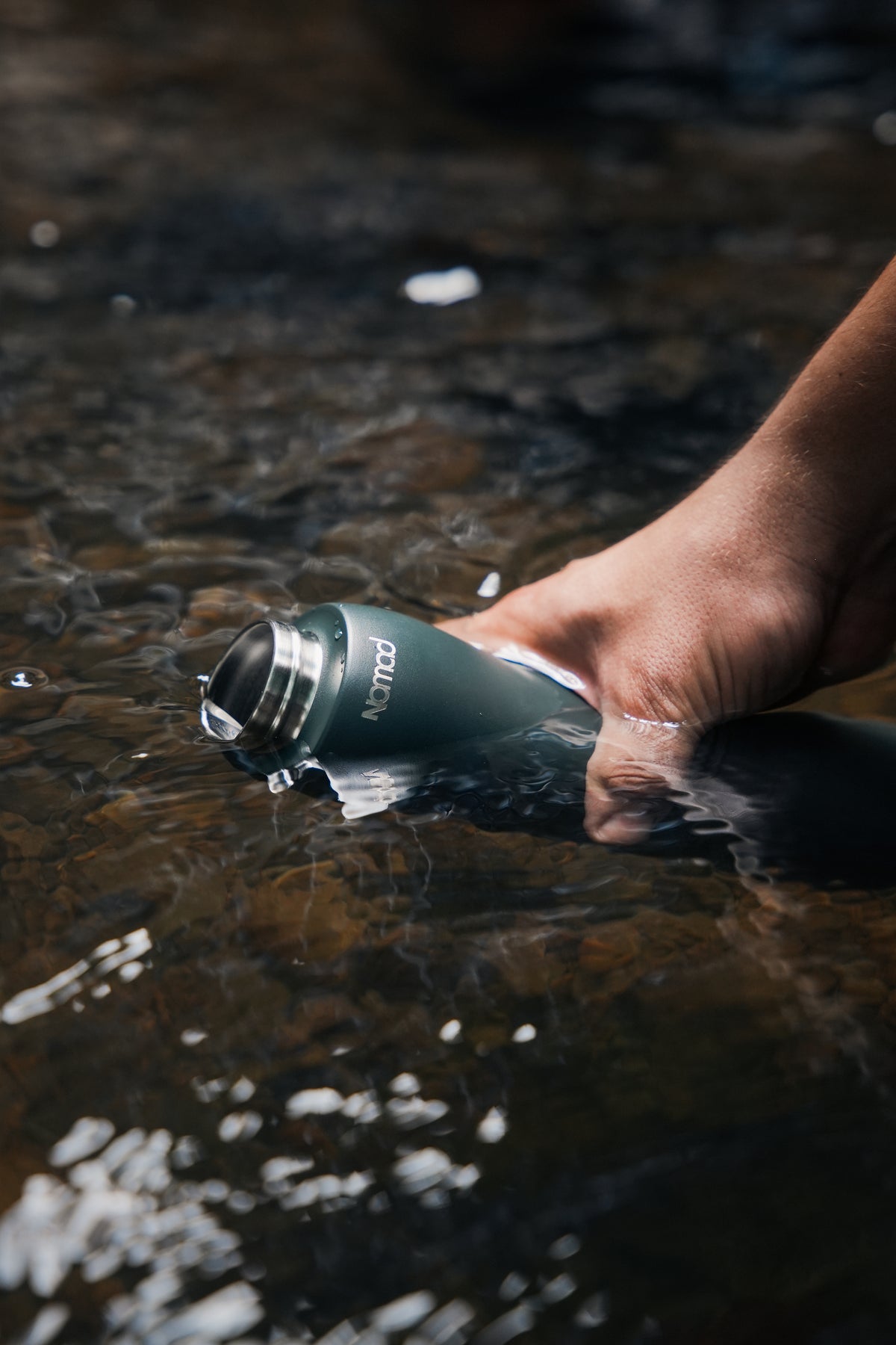 A hand holding a NOMAD SafeSip water purifier bottle being refilled from a clear stream during an outdoor hike.