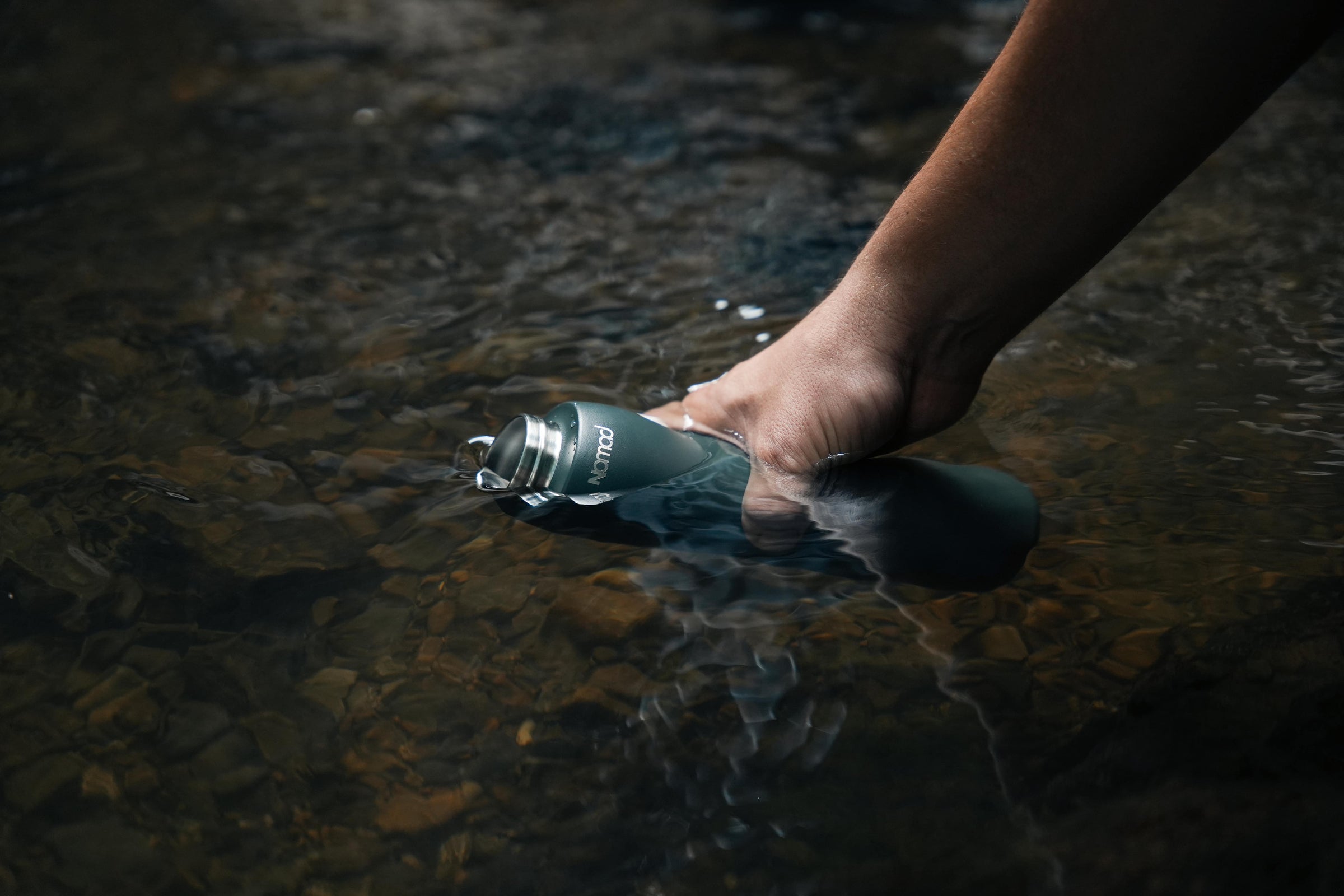 A person's hand holding a NOMAD SafeSip dark gray insulated water purifier bottle in shallow, clear water with rocks visible underneath.