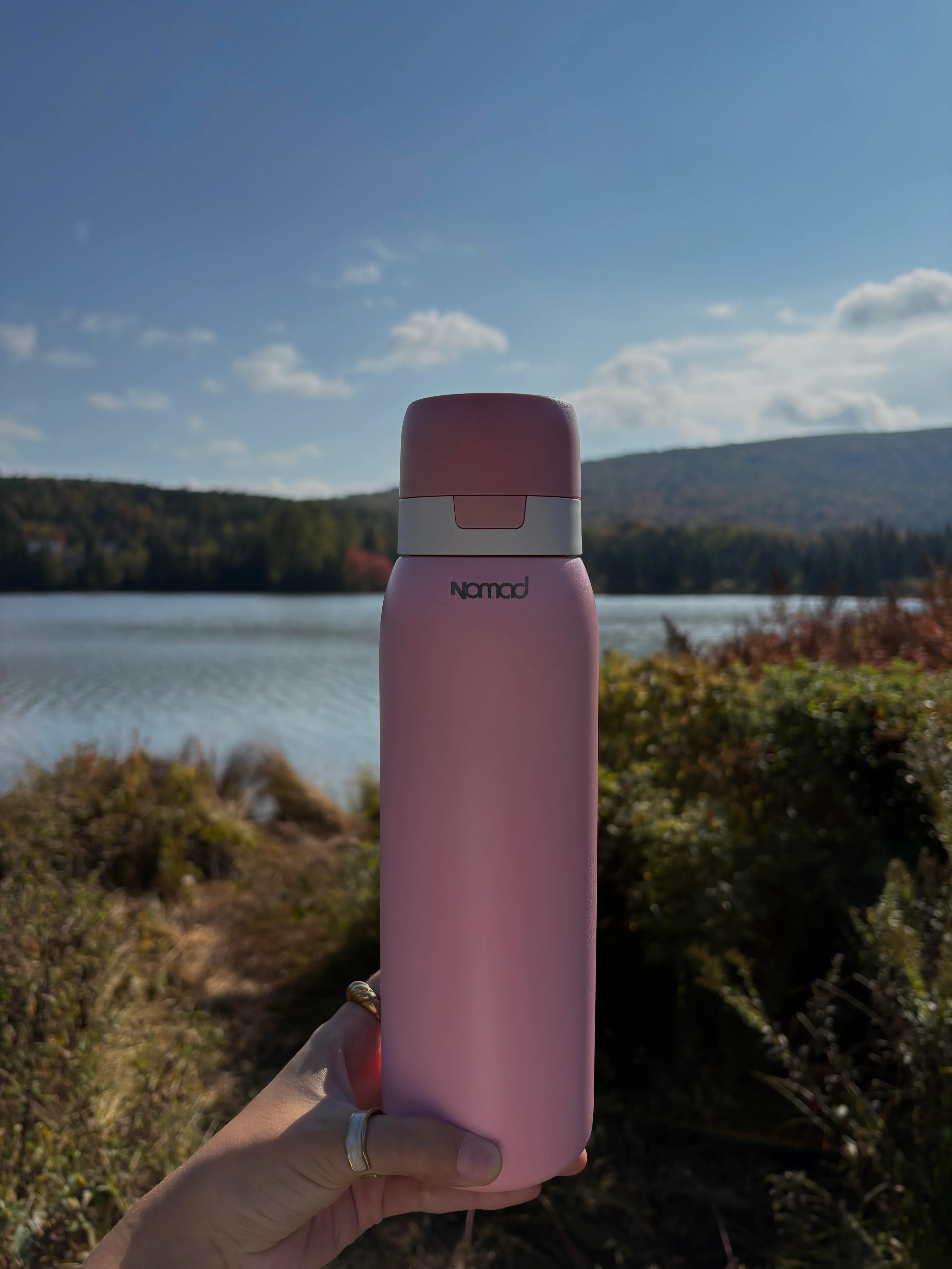 Pink SafeSip bottle held in front of a scenic lake with trees and blue sky.