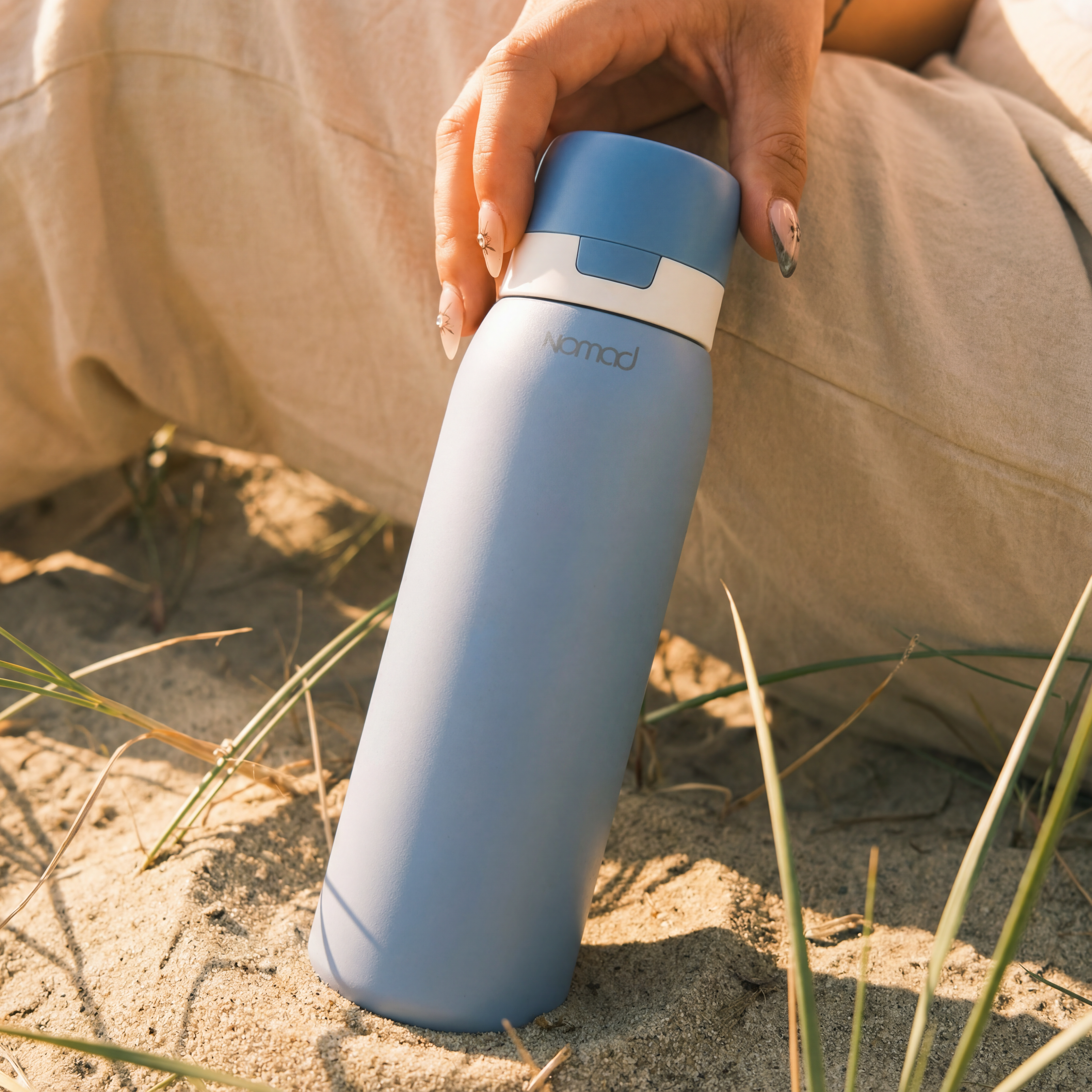 SafeSip water bottle hold by a hand outside on the sand, close to the sea. 