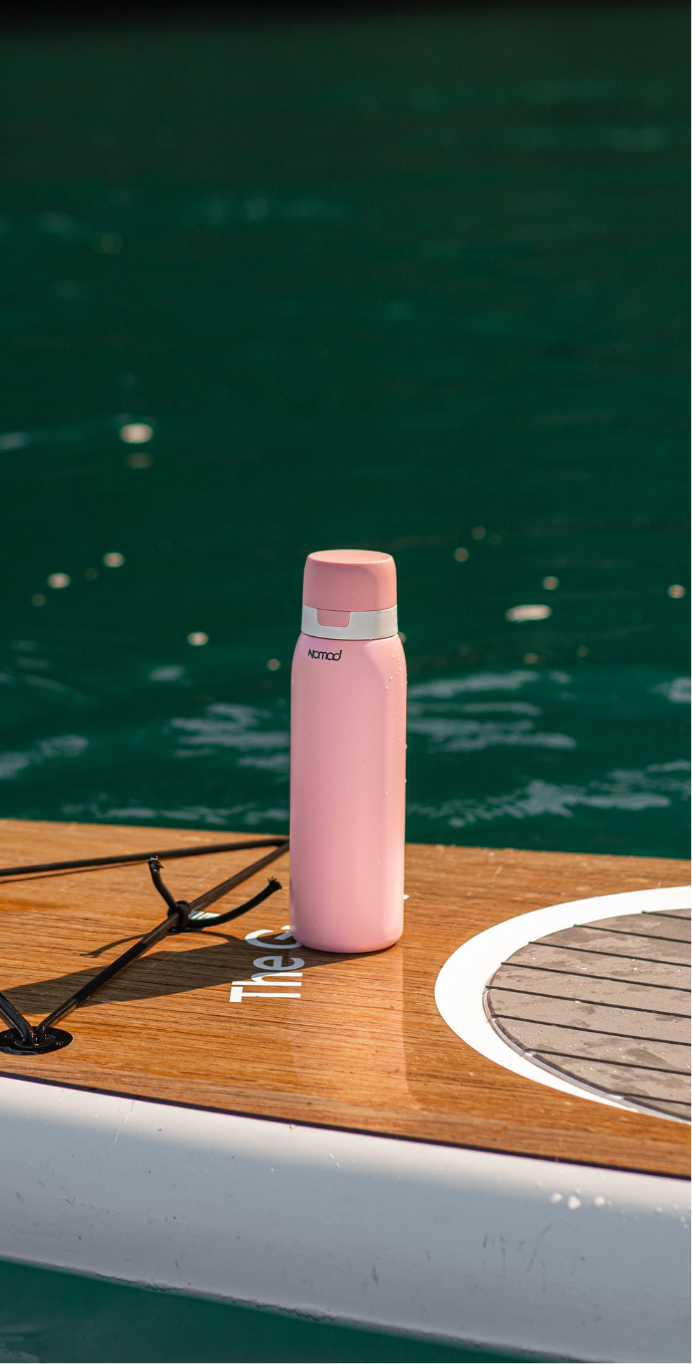 A pink stainless steel vacuum-insulated NIOMAD SafeSip water purifier bottle with a white lid sits on a wooden paddle board deck with green water in the background.