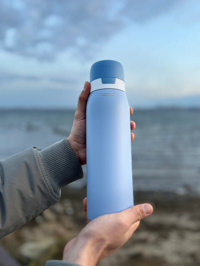 Person holding a NOMAD blue insulated bottle by a body of water. 